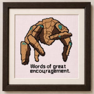 May include: A framed cross-stitch embroidery featuring a tan and brown stone-like creature with teal accents, positioned above the black text "Words of great encouragement." The artwork is enclosed in a dark brown wooden frame with a white mat.