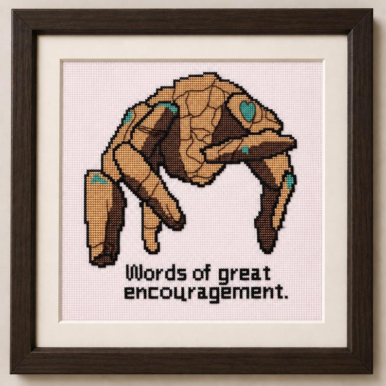 May include: A framed cross-stitch embroidery featuring a tan and brown stone-like creature with teal accents, positioned above the black text "Words of great encouragement." The artwork is enclosed in a dark brown wooden frame with a white mat.