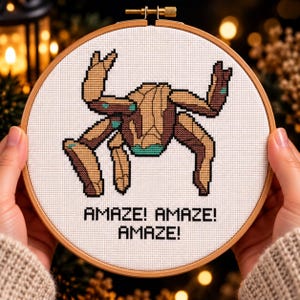 May include: A cross-stitch embroidery in a round wooden hoop featuring a brown and tan segmented creature with teal accents on a white fabric background. The text AMAZE! AMAZE! AMAZE! is stitched in black capital letters below the creature design.