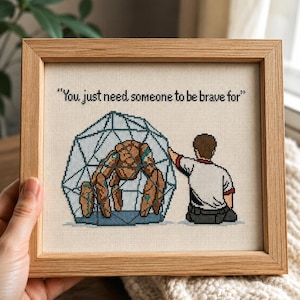 Puede incluir: Obra de arte de punto de cruz en un marco de madera. El diseño presenta una jaula geométrica que contiene una criatura marrón, con una persona que se extiende. El texto dice: "You just need someone to be brave for."
