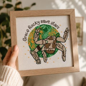 Puede incluir: Arte de punto de cruz en un marco de madera clara. El diseño presenta una figura robótica en tonos marrones y beige, sobre un globo verde y amarillo. El texto "Grace Rocky save stars" está arqueado alrededor del globo.