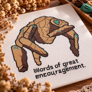 May include: A cross-stitch embroidery on white fabric featuring a stylized brown and tan rock-like creature with teal accents. Below the creature, the text reads, "Words of great encouragement." The design uses a grid-like stitch pattern.