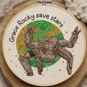 Puede incluir: Arte bordado en un aro de madera. El diseño presenta una figura marrón con forma de árbol con los brazos extendidos, sobre un fondo verde y naranja. El texto "Grace Rocky save stars" está bordado alrededor del borde.