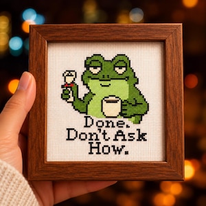 以下が含まれることがあります： ダークブラウンの木製フレームに入った正方形のクロスステッチ刺繍。白いマグカップと白い花を持った緑色のカエルが描かれています。カエルの下には「Done. Don't Ask How.」という文字が白い布地に刺繍されています。