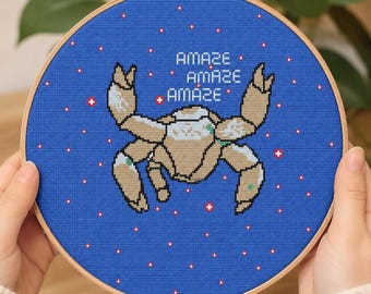 May include: A circular wooden embroidery hoop containing blue fabric with a cross-stitched tan crab in the center. The text "AMAZE AMAZE AMAZE" is stitched in white above the crab, surrounded by small white and red star-like accents.