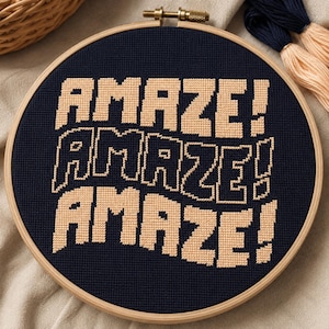 May include: A cross-stitch embroidery hoop with the word "AMAZE!" in a repeating pattern. The text is in a light tan color against a dark navy blue background. The wooden hoop is visible, and embroidery floss is in the background.