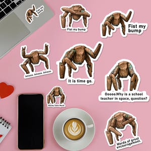Può includere: Una collezione di adesivi con una creatura marrone dall'aspetto robotico in varie pose. Alcuni adesivi hanno testo come "Fist my bump", "Amaze. Amaze. Amaze." e "It is time go." Gli adesivi sono su una superficie rosa con un laptop, un telefono e un caffè.