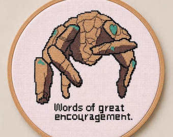 May include: A circular wooden embroidery hoop containing a cross-stitch design on light pink fabric. The design features a tan and brown rock-like creature with teal accents and the black text "Words of great encouragement." centered below.
