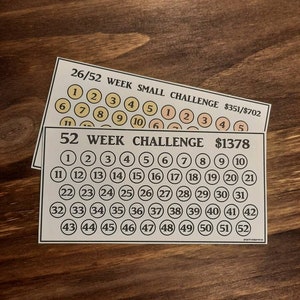 May include: Two white cards with black text and numbered circles for a savings challenge. One card is labeled "26/52 WEEK SMALL CHALLENGE" and the other "52 WEEK CHALLENGE". The cards are on a dark wood surface.