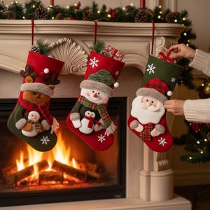 May include: Three Christmas stockings hang above a fireplace. Each stocking is red and green, with festive designs including a reindeer, snowman, and Santa Claus. White snowflakes are embroidered on the stockings. A person is holding the Santa stocking.