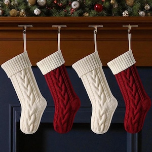 Peut inclure: Quatre bas de Noël en tricot torsadé suspendus à une cheminée. Deux sont de couleur crème et deux sont rouges. Chaque bas a un revers crème et une boucle pour l'accrocher. La cheminée est en bois foncé, avec des décorations de Noël en arrière-plan.