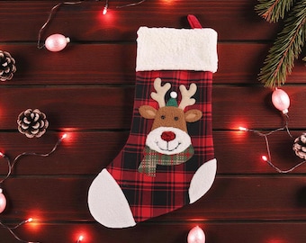 Pode incluir: Meia de Natal com um design de rena. A meia é xadrez vermelha e preta com punho e biqueira brancos. A rena tem um rosto castanho, chifres e um cachecol verde. Pinhas e luzes circundam a meia.