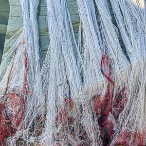 Puede incluir: Primer plano de redes de pesca, con redes blancas y rojas. Las redes están drapeadas sobre una superficie de madera, con la red blanca cayendo en cascada. Se ven cuerdas y flotadores rojos y amarillos, creando un efecto texturizado y en capas.