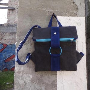 Puede incluir: Mochila negra con detalles azules, colgada en una pared de hormigón. La mochila tiene una cremallera azul, correas y un gran anillo azul. El fondo incluye una pared de ladrillo con graffiti.