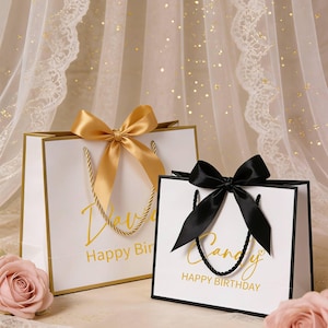 May include: Two white gift bags with gold trim and handles, each with a satin bow. One bag has a gold bow and the text "Happy Birthday" in gold script. The other bag has a black bow and the word "Candy" and "Happy Birthday" in gold script.