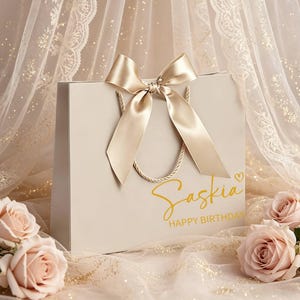 May include: A cream-coloured gift bag with a gold satin bow and rope handles. The bag features the name "Saskia" and "Happy Birthday" in gold script. Soft pink roses and a sheer, lacy fabric with gold flecks surround the bag.