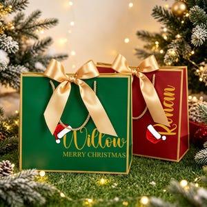 Op de afbeelding: Twee rechthoekige cadeauzakjes, een groene en een rode, elk voorzien van een gouden satijnen strik, gouden koordhengsels en een kleine kerstmuts-afbeelding. De groene tas toont de naam Willow en de tekst MERRY CHRISTMAS in gouden letters.