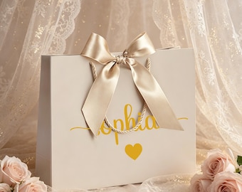 May include: A rectangular off-white gift bag featuring the name Sophia in yellow cursive script with a yellow heart below it. The bag is adorned with a large, centred champagne-coloured satin ribbon bow tied around the top handles.