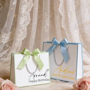 May include: Two white gift bags with pastel green and blue accents. Each bag has a matching ribbon bow and silver rope handles. One bag says "Arnaud Happy Birthday" and the other says "Andrea Happy Birthday".