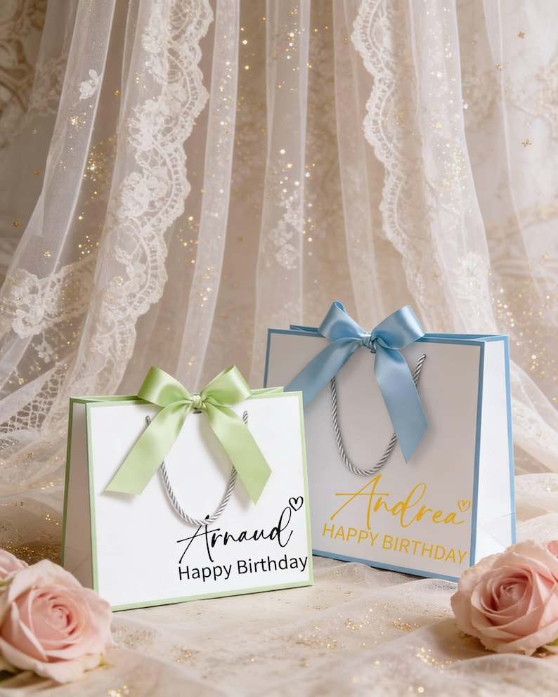 May include: Two white gift bags with pastel green and blue accents. Each bag has a matching ribbon bow and silver rope handles. One bag says "Arnaud Happy Birthday" and the other says "Andrea Happy Birthday".