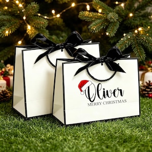Op de afbeelding: Twee witte cadeauzakjes met zwarte randen en zwarte satijnen linthengsels gestrikt tot strikken. Het voorste zakje toont de naam Oliver in zwarte schrijfletters, een rood-witte kerstmuts en de tekst MERRY CHRISTMAS in zwarte hoofdletters.