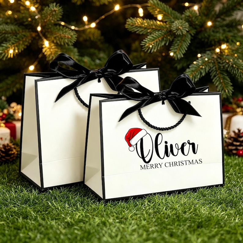 Op de afbeelding: Twee witte cadeauzakjes met zwarte randen en zwarte satijnen linthengsels gestrikt tot strikken. Het voorste zakje toont de naam Oliver in zwarte schrijfletters, een rood-witte kerstmuts en de tekst MERRY CHRISTMAS in zwarte hoofdletters.