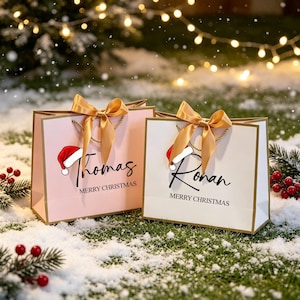 Op de afbeelding: Twee rechthoekige cadeauzakjes, een lichtroze en een witte, elk voorzien van een gouden strik, een rood-witte kerstmuts en de tekst Merry Christmas met de namen Thomas op het roze zakje en Ronan op het witte zakje.