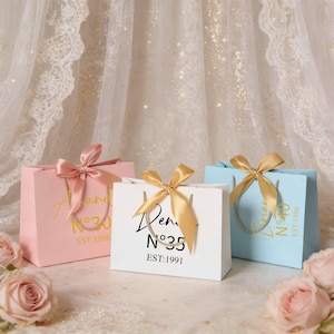 May include: Three gift bags in pink, white, and light blue, each with a satin ribbon bow and rope handles. The bags have gold text and numbers, with the text "an", "Dem", and "N&deg;40" visible. The text "EST:1996", "N&deg;35 EST:1991", and "EST:1986" are also visible.