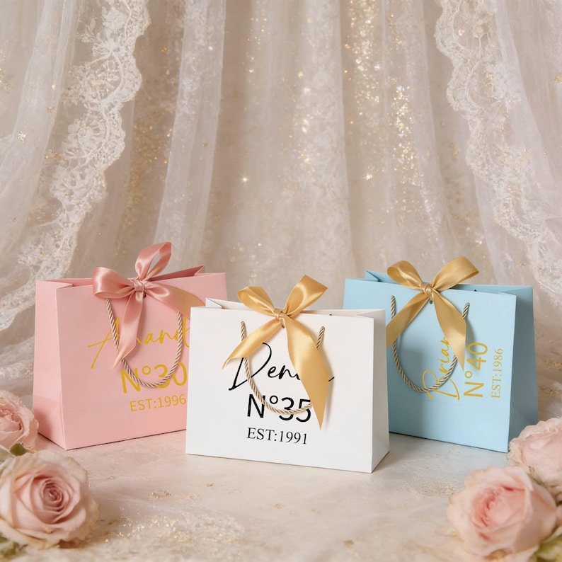 May include: Three gift bags in pink, white, and light blue, each with a satin ribbon bow and rope handles. The bags have gold text and numbers, with the text "an", "Dem", and "N&deg;40" visible. The text "EST:1996", "N&deg;35 EST:1991", and "EST:1986" are also visible.