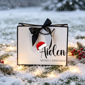 Op de afbeelding: Een witte cadeauzak met zwarte randen en een zwarte satijnen strik. De voorzijde toont de naam Aiden in zwart cursief schrift, een afbeelding van een rood-witte kerstmuts en de tekst MERRY CHRISTMAS in zwarte hoofdletters onder de naam.