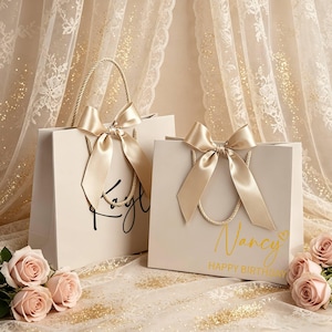May include: Two cream gift bags with gold satin bows and rope handles. One bag has the name "Faye" in black script, the other says "Nancy HAPPY BIRTHDAY" in gold. Pink roses are in the foreground, with a sheer, gold-speckled curtain backdrop.