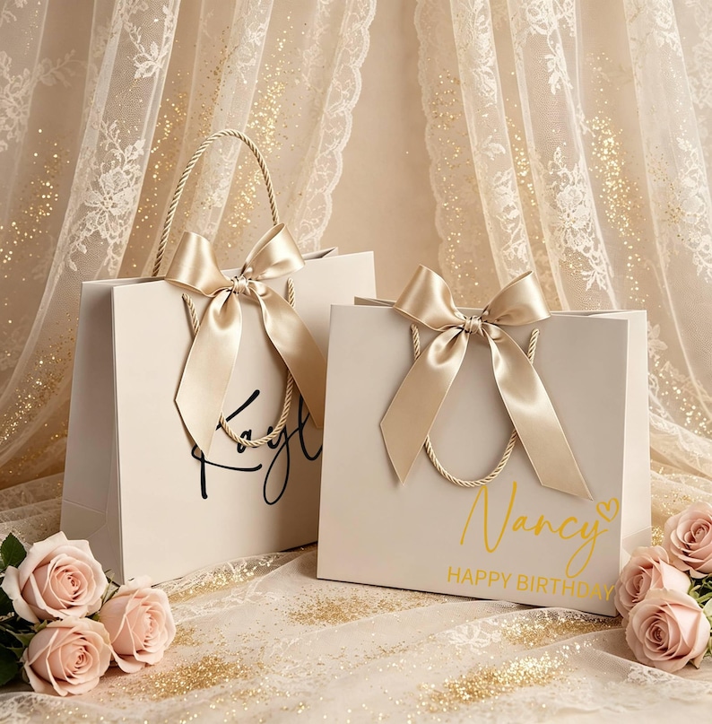 May include: Two cream gift bags with gold satin bows and rope handles. One bag has the name "Faye" in black script, the other says "Nancy HAPPY BIRTHDAY" in gold. Pink roses are in the foreground, with a sheer, gold-speckled curtain backdrop.