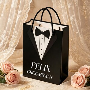 May include: A black gift bag featuring a white tuxedo shirt front graphic with a black bow tie and three buttons. The text FELIX GROOMSMAN is printed in white on the front. The bag has two black twisted rope handles and a white interior lining.
