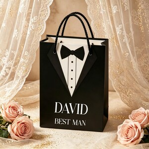 May include: A black gift bag featuring a white tuxedo shirt graphic with a black bow tie. The bag has black rope handles and the white text DAVID BEST MAN printed on the front. The design mimics formal attire with a black lapel and white shirt front.