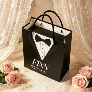 May include: A black gift bag with a white tuxedo shirt and black bow tie graphic. The text FINN BEST MAN is printed in white on the front. The bag features two black twisted rope handles and a white interior lining, standing upright.