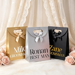May include: Three gift bags in gold, silver, and black with die-cut handles and satin bows. The gold bag features white text reading Milo GROOMSMAN, the silver bag has black text reading Ronan BEST MAN, and the black bag has gold text reading Zane GROOMSMAN.