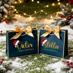 Op de afbeelding: Twee donkerblauwe cadeauzakjes met een gouden rand en een grote gouden satijnen strik aan de bovenkant. Elke tas is voorzien van een rood-wit kerstmutspictogram, de naam Victor of Stella in gouden letters en de tekst MERRY CHRISTMAS.