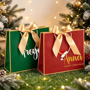 Op de afbeelding: Twee rechthoekige cadeauzakjes, &eacute;&eacute;n donkergroen en &eacute;&eacute;n dieprood, elk met gouden randen, gouden koordhengsels en een grote gouden satijnen strik. De rode tas toont de naam Anna in gouden schrift met een kerstmuts en de tekst Merry Christmas.