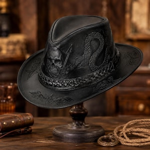 Könnte beinhalten: Ein schwarzer Fedora-Hut mit geprägten Details eines Totenkopfes und einer Schlange auf der Krone. Der Hut verfügt über ein geflochtenes Lederband an der Basis und aufwendige florale Schnitzereien auf der breiten Krempe in matter Optik.