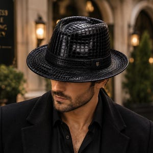 Può includere: Un cappello fedora nero con un motivo testurizzato in pelle di coccodrillo lucida. Il cappello presenta una corona strutturata, una tesa larga e una fascia nera attorno alla base con un piccolo emblema metallico sul lato. Il materiale è riflettente.