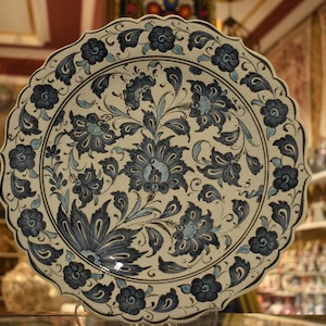 May include: A decorative ceramic plate with a cream-colored base and intricate blue floral patterns. The plate has a scalloped edge and is likely handmade, showcasing a traditional design. The floral design includes stylized flowers and leaves.