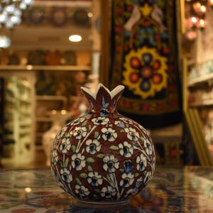 May include: A decorative ceramic pomegranate-shaped vase. The vase is primarily brown with a floral pattern of white and blue flowers. The top of the vase has a crown-like design. The background shows a shop with shelves of pottery.