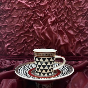 Op de afbeelding: Een decoratieve koffiekop en schotel set. De kop heeft een zwart-wit geometrisch driehoekpatroon met een goud-bruine rand. De schotel heeft een bijpassend ontwerp met een rode accent.