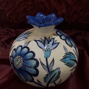 May include: A ceramic vase with a round body and a blue flower-shaped top. The vase is white with a textured surface and features blue floral designs. The flowers and leaves are painted in shades of blue.