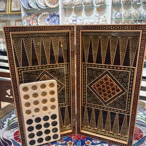 Puede incluir: Un tablero de backgammon abierto con un soporte de piezas de juego color crema. El tablero es marrón con intrincados diseños geométricos en negro, dorado y rojo. El soporte de piezas de juego tiene piezas de juego negras y crema. El fondo incluye platos y cerámica decorativos.