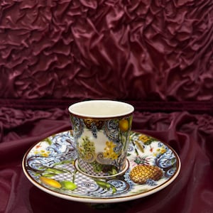 Op de afbeelding: Een decoratieve kop en schotel set. De kop en schotel hebben een gedetailleerd bloemen- en fruitontwerp met gele, groene en blauwe accenten. De set staat op een bordeauxrode stoffen achtergrond.