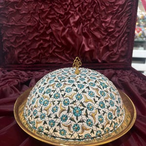 Puede incluir: Un plato de servir decorativo con tapa abovedada, con un estampado floral en turquesa y dorado sobre fondo blanco. El plato se asienta sobre una base dorada, con un remate dorado a juego. El fondo es de tela roja oscura.