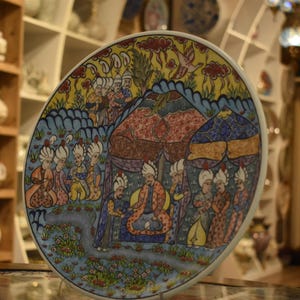 Puede incluir: Un plato de cerámica decorativo con una ilustración detallada y colorida. La obra de arte representa figuras con ropa ornamentada bajo tiendas de campaña, con un cielo azul y amarillo. El plato es redondo y probablemente pintado a mano, mostrando un diseño tradicional.