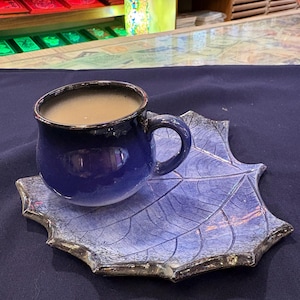 Op de afbeelding: Een blauwe keramische koffiekop met een donkere rand staat op een bijpassende blad-vormige schotel. De kop bevat een lichtbruine vloeistof. De schotel heeft een gedetailleerd bladnervenpatroon en een donkere rand. De set staat op een donkerblauwe ondergrond.