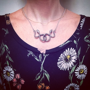 May include: A silver snake necklace with two intertwined snakes. The necklace is worn by a person with a black floral patterned shirt.